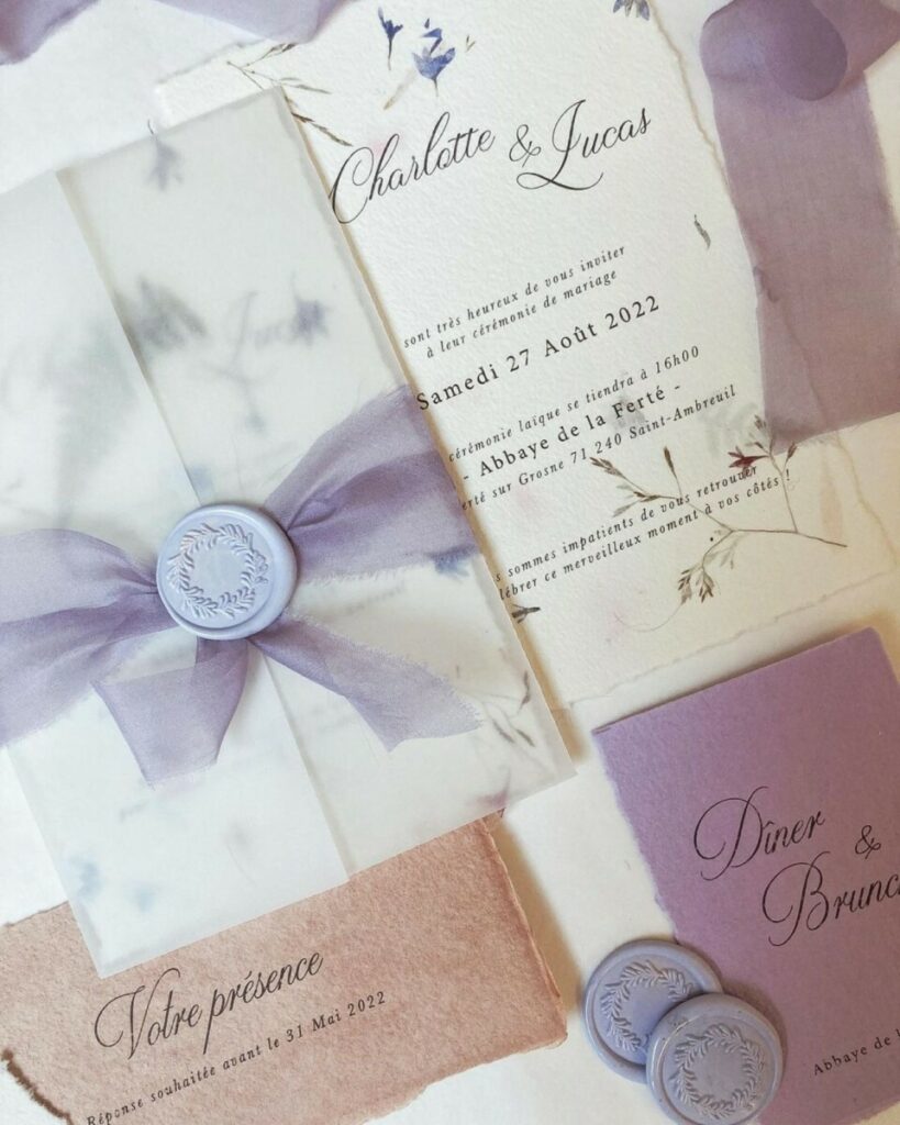 Faire-part de mariage avec papier fait main et inclusions de fleurs des champs – cachet de cire lavande – ruban mauve et pochette calque