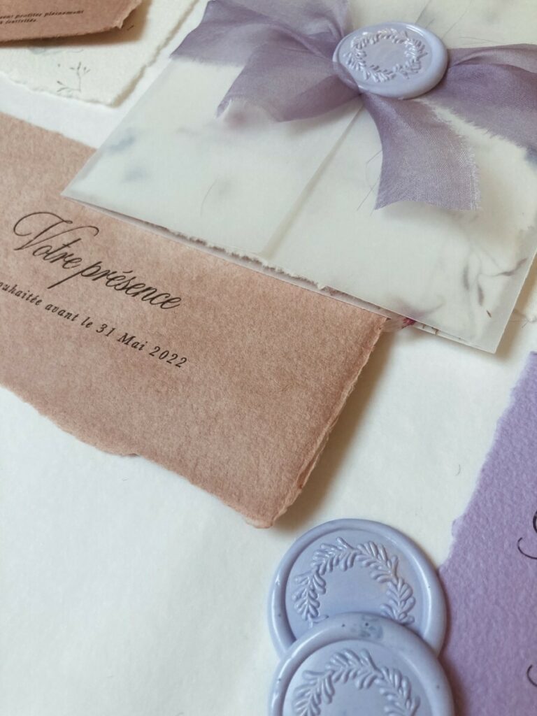 Faire-part de mariage avec papier fait main et inclusions de fleurs des champs – cachet de cire lavande – ruban mauve et pochette calque